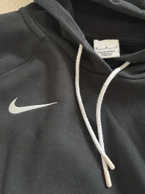 Svart Nike hoodie med vit logga - Snygg svart hoodie från Nike med vit broderad Swoosh-logga på bröstet och vita dragsnören i huvan. Klassisk känguruficka framtill och mjukt material som känns skönt mot huden. Perfekt för en avslappnad och sportig stil.