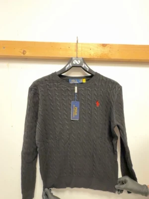 Svart kabelstickad tröja Polo Ralph Lauren - Snygg svart kabelstickad tröja från Polo Ralph Lauren med klassisk ranges röd logga broderad på bröstet. null har rund hals double ribbade muddar vid ärmslut och nederkant. Perfekt för dig som gillar tidlös och stilren design.