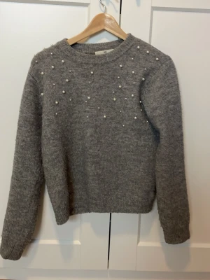 Grå stickad tröja med pärlor - Snygg grå stickad tröja med pärldetaljer på axlar och bröst. Tröjan har rund halsringning, långa ärmar och ribbstickade muddar. Enkel och stilren design. Storlek 146/152 lite luddig.