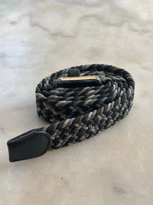 Flätat bälte - Säljer ett snyggt flätat bälte i blått och grått med silverfärgat spänne. Bältet har en flexibel design och är tillverkat av syntetmaterial med detaljer i skinn. Bältet är 107cm långt. 