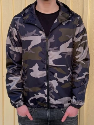 Camo windbreaker - Snygg vindjacka i camouflagemönster med dragkedja framtill och huva. Jackan har elastiska muddar vid ärmsluten och är tillverkad i ett lätt, vindtätt material. Perfekt för dig som gillar streetwear och vill ha en stilren look.
