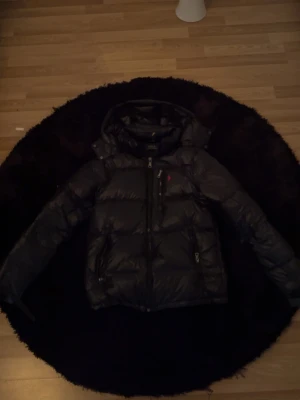 Svart dunjacka från Polo Ralph Lauren - Snygg svart dunjacka från Polo Ralph Lauren med huva och dragkedja framtill. Jackan har två sidofickor med dragkedja och en liten röd logga på bröstet. Perfekt för kalla vinterdagar och riktigt stilren look.