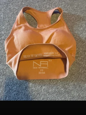 Rostfärgad ribbad sport-bh XS - Säljer en ribbad sport-bh från New Apparel i en snygg rostfärgad ton. Bh:n har brottarrygg och bred resår under bysten för extra stöd. Tillverkad i ett stretchigt material som består av 90% polyamid och 10% elastan. Storlek XS (32/34).