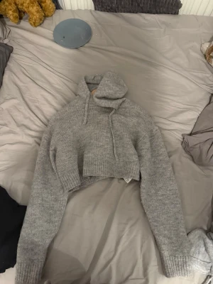 Grå cropped hoodie från Primark - Säljer en grå stickad hoodie från Primark i storlek XS. Tröjan är cropped med långa ärmar, ribbstickade muddar och huva med dragsnören. Perfekt för dig som gillar en avslappnad och trendig look. Materialet är mjukt och värmande.