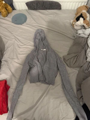 Grå croppad kofta med dragkedja - Säljer en grå croppad kofta från Gina  med dragkedja framtill och långa ärmar. Koftan är stickad i mjukt material och har ribbade muddar vid nederkant och ärmslut. Perfekt för en chill och avslappnad stil.