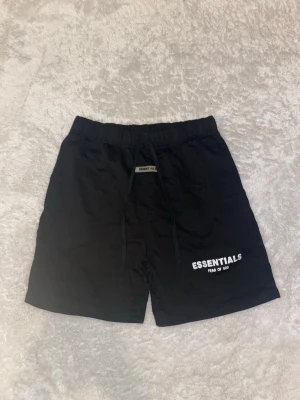 Svarta Essentials shorts med snörning - Svarta shorts från Essentials Fear of God med snörning i midjan och logotyp tryckt på framsidan. De har en enkel och clean design, perfekt för en avslappnad look. Materialet känns mjukt och bekvämt, och passformen är normal.