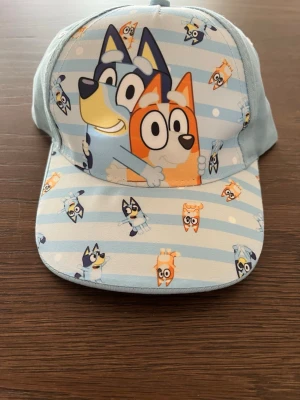 Blå keps med Bluey-motiv - Säljer en ljusblå keps med motiv från Bluey och Bingo på framsidan. Kepsen har ett randigt mönster i vitt och blått samt flera små figurer från serien. Justerbar baktill och tillverkad i bomullsmaterial. Perfekt för dig som gillar Bluey!