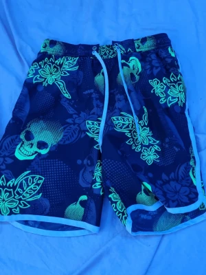 Svarta badbyxor med dödskallar KappAhl - Coola svarta badbyxor från KappAhl med neongröna dödskallar och bladmönster. De har vit dragsko i midjan, vita kanter och meshfoder på insidan. Perfekta för stranden eller poolen för dig som vill sticka ut.