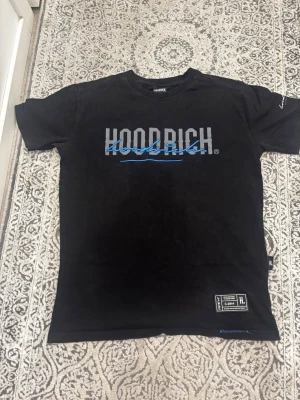 Svart Hoodrich t-shirt med tryck - Svart t-shirt från Hoodrich med stort tryck i vitt och blått på bröstet. Klassisk rund hals och korta ärmar. Tillverkad i mjuk bomull och har en liten etikett nertill. Perfekt för dig som gillar streetwear och vill ha en clean look.