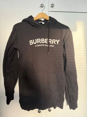 Svart Burberry hoodie med tryck - Svart hoodie från Burberry med vit text 'BURBERRY LONDON ENGLAND' på bröstet. Klassisk modell med huva och känguruficka framtill. Mjuk bomullskänsla och ribbade muddar vid ärmslut och nederkant. Perfekt för en stilren och trendig look.