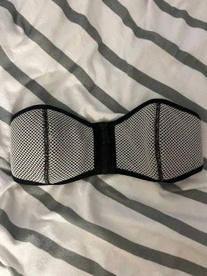 TRIANGL bikinitopp - Säljer en snygg bandeau bikiniöverdel i svartvitt meshmönster. Överdelen har dragkedja framtill och breda kanter för extra stöd. Perfekt för dig som vill ha en stilren och sportig look på stranden. Från triangl i storlek S men passar snarare XS🥰