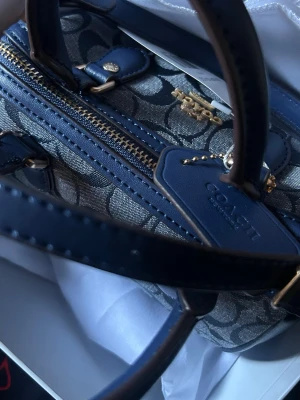 Blå handväska från Coach - Snygg blå handväska från Coach med klassiskt monogrammönster i canvas och detaljer i mörkblått läder. Väskan har guldfärgade metalldetaljer, dragkedja och Coach-tag. Perfekt för dig som vill ha en trendig och stilren accessoar.