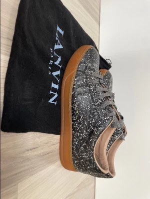 Lanvin svarta sneakers med mönster - Snygga svarta sneakers från Lanvin med vitt stänkmönster och bruna gummisulor. Skorna har grå snörning och är tillverkade i läder med en låg siluett. Perfekta för dig som vill sticka ut med en unik design och exklusiv känsla. Lanvin läderskor i mycket gott skick, dammpåse finns. !!! AUTENTISK Storlek 8 / verklig storlek EU 43 /. Nypris ca 6000-8000 kr  Meddela mig för mer information, kan skicka mer bilder och pris kan diskuteras!