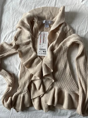 Beige volangkofta från MISU Knitwear - Supersöt beige kofta från new yorker  med volangkant längs hela öppningen och nederkanten. djup v-ringning. Oanvänd beige volangtopp med ihåliga mönster. Bekväm och kortare framtill. Säljer även samma modell i svart (kontakta för paketpris)