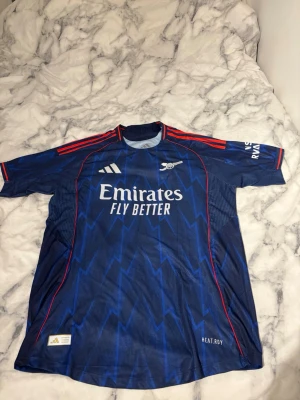 Arsenal bortatröja Adidas blå - Snygg Arsenal bortatröja från Adidas i mörkblått med blixtmönster och röda detaljer på axlar och ärmslut. Tröjan har Emirates Fly Better-tryck på bröstet och är tillverkad i lätt, ventilerande material. Perfekt för fotboll eller träning.