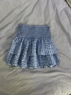 Blommig volangkjol i blått - Supersöt kort kjol med blått blommönster och dubbla volanger. Kjolen har en smockad midja som ger en bekväm och stretchig passform. Perfekt för dig som gillar en lekfull och trendig stil. Materialet känns lätt och luftigt.