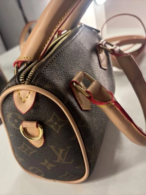 monogram handväska - Snygg handväska  i klassiskt brunt monogrammönster med ljusa läderdetaljer och guldiga metalldelar. Väskan har både handtag och axelrem samt dragkedja upptill. Perfekt accessoar för dig som gillar lyxig och tidlös design.