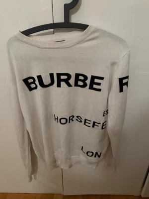 Vit stickad tröja från Burberry - Vit stickad tröja från Burberry med svart textmönster över hela plagget. Tröjan har rund halsringning och långa ärmar. Materialet är mjukt och tunt, perfekt för lager-på-lager och en cool streetstyle-look.