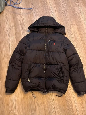 Svart pufferjacka från Polo Ralph Lauren - Snygg svart pufferjacka från Polo Ralph Lauren med huva och dragkedja framtill. Jackan har två dragkedjeförsedda fickor och den klassiska röda loggan på bröstet. Perfekt för kalla vinterdagar och riktigt skön att bära.