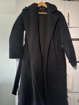 Svart teddy-kappa  - Lång svart teddy-kappa från NEW YORKER  med bälte i midjan och stora fickor. Perfekt för kalla dagar när du vill vara både snygg och bekväm. Storlek 34