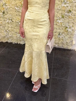 Ljusgul långklänning med rosett - Elegant ljusgul långklänning i glansigt tyg med subtilt blommönster. Klänningen är axelbandslös, har snörning i ryggen och en stor dekorativ rosett baktill. Figurnära passform som framhäver siluetten, perfekt för speciella tillfällen.