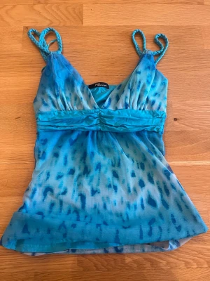 Jane Norman Blå mönstrad linnetopp med knyt  - Säljer en blå linnetopp från Jane norman med fläckigt mönster i olika blå nyanser. Toppen har smala, tvinnade axelband, v-ringning och ett brett band under bysten som knyts i ryggen. Materialet är tunt och lätt, perfekt för varma dagar.