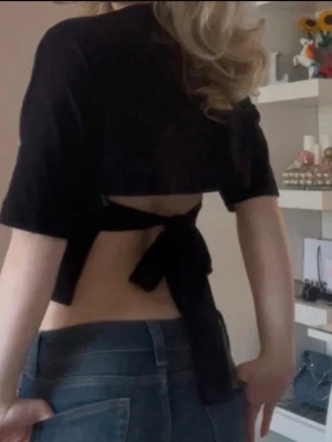 Svart croppad topp med knytband - Trendig svart croppad topp med korta ärmar och långa knytband i midjan som ger en snygg öppen rygg. Köpt från Nelly, använd fåtal gånger.