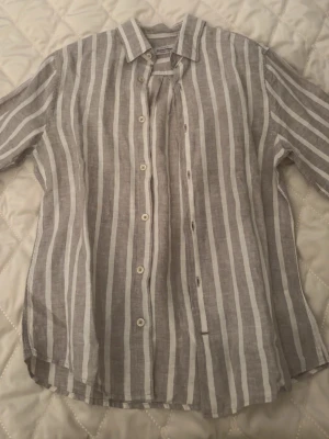 Randig linneskjorta i beige och vit - Säljer en luftig skjorta i linne med vertikala beige och vita ränder. Skjortan har klassisk krage, knappar framtill och långa ärmar. Perfekt för en avslappnad och stilren look.