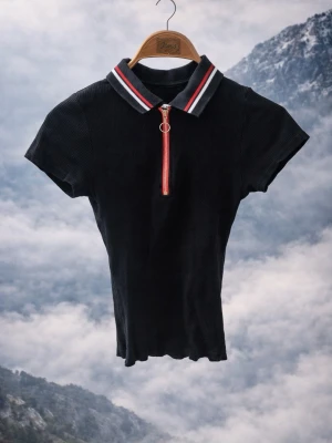 Svart ribbad polotopp med dragkedja - Snygg svart ribbad polotopp med korta ärmar och krage med röd, vit och svart randig detalj. Dragkedja framtill med ringdetalj ger en sportig touch. Perfekt till jeans eller kjol för en trendig look.