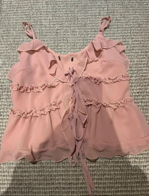 Rosa linnetopp med volanger och snörning - Supersöt rosa linnetopp med volangdetaljer och snörning framtill. Toppen har tunna axelband och är tillverkad i ett lätt, luftigt material som passar perfekt till varma dagar. Perfekt för dig som gillar romantisk och feminin stil. Aldrig använd och har reglerbara band