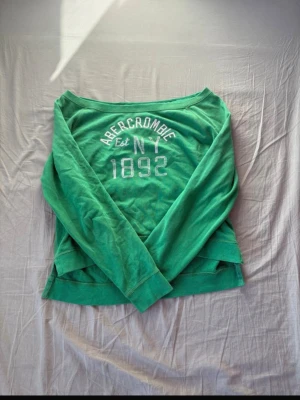 Grön sweatshirt från Abercrombie - Cool grön sweatshirt från Abercrombie med tryck 'Abercrombie Est NY 1892' i vitt på bröstet. Tröjan har rund halsringning, långa ärmar och är gjord i mjukt bomullsmaterial. Perfekt för en avslappnad och sportig look.