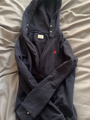 Svart hoodie från US Polo Assn - Mörkblå hoodie från US Polo Assn med huva och dragkedja. Diskret röd logga broderad på bröstet och metallöljetter vid snörning. Mjukt material som känns skönt mot huden och klassisk design med ribbade muddar. Perfekt för en avslappnad look. Funkar på både tjejer och killar. Jag brukar ha xs o den funkar för mig😅