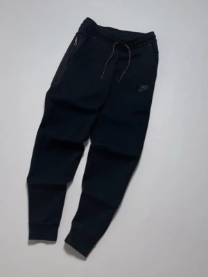 Svarta Nike mjukisbyxor med dragsko - Säljer ett par svarta mjukisbyxor från Nike med dragsko i midjan och ribbade muddar vid bensluten. Byxorna har fickor med dragkedja och en diskret Nike-logga på låret. Perfekta för chill dagar eller träning. Tillverkade i mjukt och bekvämt material.