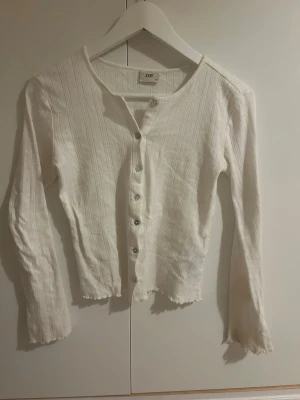 Vit ribbad kofta från JDY - Säljer en vit ribbad kofta från JDY i storlek XS. Koftan har långa ärmar, knappar framtill och ett diskret mönster i tyget. Den har en croppad passform och vågig kant nedtill för en trendig look.