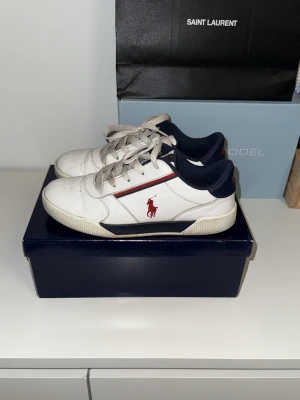Vita sneakers från Polo Ralph Lauren - Snygga vita sneakers från Polo Ralph Lauren med marinblå detaljer och röd logga på sidan. Klassisk låg modell med vita snören och perforerad tå. Skorna har en sportig vibe och passar perfekt till jeans eller shorts. Obs!! Lådan tillkommer inte då den slängdes under flyttning.