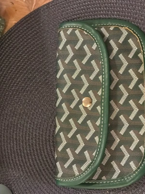 Fake Grön mönstrad plånbok med tryckknapp - Snygg plånbok i grönt med vitt grafiskt mönster och gröna kanter. Plånboken har en rundad form och stängs med en guldfärgad tryckknapp. Materialet ser ut att vara canvas med detaljer i konstläder. Perfekt för dig som gillar unika accessoarer.