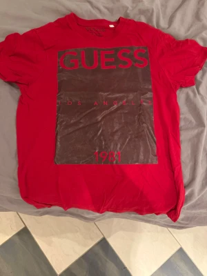 Röd GUESS t-shirt med tryck - Snygg röd t-shirt från GUESS med stort svart och rött tryck på framsidan där det står 'GUESS Los Angeles 1981'. Klassisk passform och korta ärmar. Perfekt för dig som vill ha en statement-tee med streetkänsla.