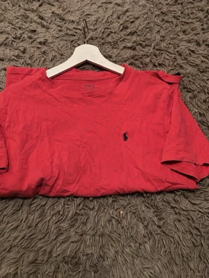 Röd t-shirt från Polo Ralph Lauren - Säljer en röd t-shirt från Polo Ralph Lauren med klassisk rund hals och den ikoniska mörkblå broderade loggan på bröstet. T-shirten är kortärmad och gjord i mjuk bomull, perfekt för en avslappnad och stilren look.