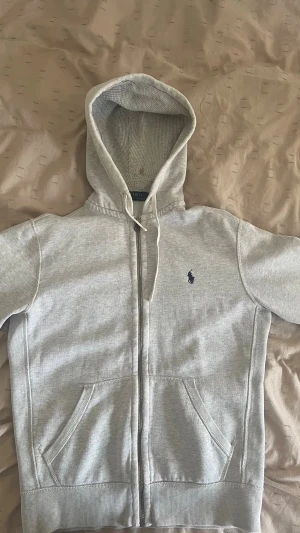 Grå hoodie från Polo Ralph Lauren - Ljusgrå hoodie från Polo Ralph Lauren med dragkedja, två fickor framtill och klassisk blå logga broderad på bröstet. Tillverkad i mjuk bomullsblandning och har en stor huva med dragsko. Perfekt för en avslappnad och stilren look. Den har en fläck vid luvan men har varit betydligt större innan tvättning så den lär gå bort, även lite slitage där med! 