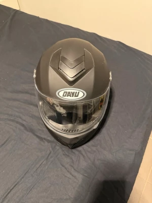 Svart integralhjälm från DAYU - Snygg svart integralhjälm från DAYU med matt finish och klart visir. Hjälmen har ventiler upptill och vadderad insida med meshfoder för extra komfort. Justerbart hakspänne med snabbfäste. Perfekt för dig som vill ha en stilren och skyddande hjälm.