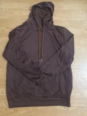 Brun hoodie med huva och dragsko - En enkel och stilren brun hoodie med huva och matchande dragsko. Hoodien har långa ärmar, ribbade muddar och en klassisk passform. Perfekt för en avslappnad look och lätt att matcha med andra plagg. Materialet känns mjukt och bekvämt.