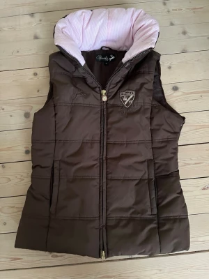 Brun dunväst från Spooks med huva - Snygg brun dunväst från Spooks med broderad logga på bröstet och guldfärgad dragkedja. Västen har en vadderad huva med ljusrosa randigt foder och två fickor framtill. Perfekt för lager på lager och riktigt skön att bära under kyliga dagar.