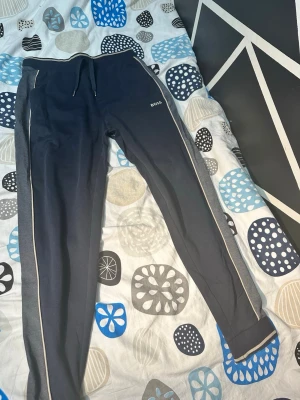 Mörkblå mjukisbyxor från Boss Storlek M - Säljer ett par mörkblå mjukisbyxor från Boss med gråa sidopaneler och vita detaljer längs benen. Byxorna har dragsko i midjan och smal passform. Boss-logga broderad på vänster lår. Perfekta för chill eller träning.