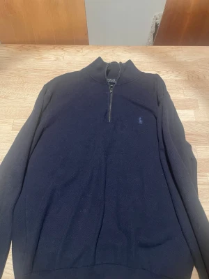 Mörkblå halvzip tröja Polo Ralph Lauren - Mörkblå stickad tröja från Polo Ralph Lauren med halvzip och hög krage. Tröjan har det klassiska broderade logot på bröstet i matchande blå ton. Perfekt för en clean och stilren look.