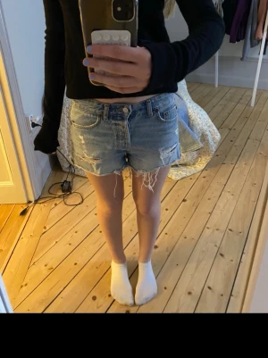 Blå denimshorts med slitningar - Säljer ett par klassiska blå jeansshorts med slitna detaljer och fransig kant. Shortsen har fem fickor, knappgylf och en avslappnad passform. Perfekta för sommardagar och enkel att matcha med olika toppar.
