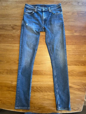 Blå slim fit jeans från Nudie - Säljer ett par klassiska blå slim fit jeans från Nudie med fem fickor och diskreta slitningar framtill. Jeansen har normal midja och är tillverkade i denim med lätt stretch för skön passform. Perfekta till en avslappnad stil.