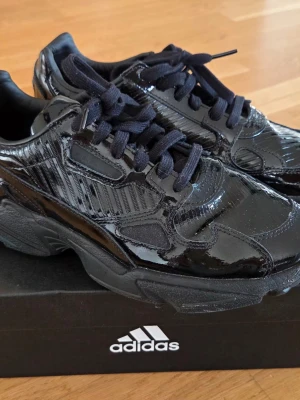 Svarta lackade sneakers från Adidas - Snygga svarta sneakers från Adidas med glansig lackad yta och svarta skosnören. Skorna har chunky sula och coola detaljer i materialet som ger en modern vibe. Perfekta för dig som vill sticka ut med stilrena men ändå unika sneakers.