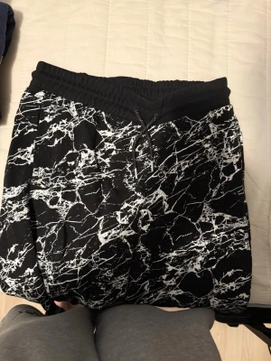 Svarta mönstrade shorts från H&M - Säljer ett par svarta shorts från H&M med vitt marmorerat mönster. De har elastisk midja med snörning och är tillverkade i mjuk bomull, perfekta för chill dagar eller träning. Snygg och avslappnad passform.