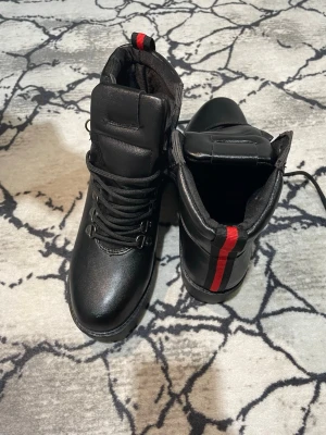 Svarta kängor med röd detalj - Snygga svarta kängor med snörning och en cool röd rand på hälen. Tillverkade i ett slätt, matt material och har vadderad plös och krage för extra komfort. Perfekta för dig som vill ha en stilren men ändå edgy look.