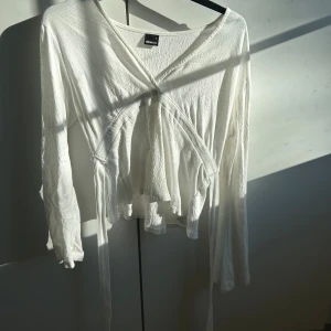 Vit omlottopp med knytband - Säljer en vit omlottopp från Gina med långa ärmar och knytband i midjan. Toppen har en v-ringad hals och är gjord i ett lätt, ribbat material.